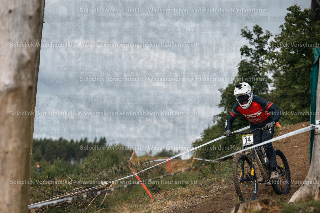 Action im Bikepark Winterberg | Ein Mountainbiker, der mit voller Geschwindigkeit einen Downhill-Kurs durchquert, dominiert das Bild. Er trägt einen weißen Helm, eine rote Jacke und schwarze Handschuhe. Sein Mountainbike ist mit gelben Felgen und einer schwarzen Rahmenfarbe ausgestattet. Der Kurs ist mit Markierungen versehen, die den Weg weisen. Im Hintergrund erstreckt sich eine dichte Waldlandschaft. Der Himmel ist bewölkt. - Realisiert mit Pictrs.com