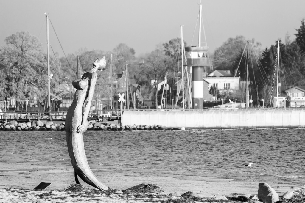Wandbild: Nixe und Leuchtturm in Eckernförde in Schwarz-Weiß | Dieses Wandbild im Querformat zeigt den Ostseestrand in Eckernförde in Schwarz-Weiß. Am Strand steht eine Nixe. Im Hintergrund kann man den Leuchtturm am Yachthafen sehen.  - Realisiert mit Pictrs.com