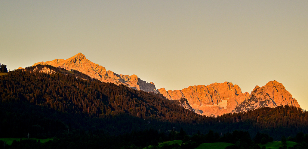 Goldenes Erwachen: Das erste Leuchten des Wettersteingebirges | Ein Anblick von magischer Stille und tiefer Erhabenheit, der den Beginn eines neuen Tages feiert: Im ersten, zarten Licht des Sonnenaufgangs erstrahlt die gesamte Kette des Wettersteingebirges mit Alpspitze und Zugspitzenmassiv in einem intensiven, warmen Alpenglühen. Die schroffen Felsgipfel leuchten wie flüssiges Gold vor dem noch blassen Morgenhimmel. Im markanten Kontrast dazu liegen die dichten, bewaldeten Berghänge im Vordergrund noch in tiefem, mystischem Schatten. Diese Aufnahme fängt die friedliche Klarheit und die berührende Herzenswärme einer erwachenden Bergwelt perfekt ein – ein Moment puren Glücks. - Realisiert mit Pictrs.com