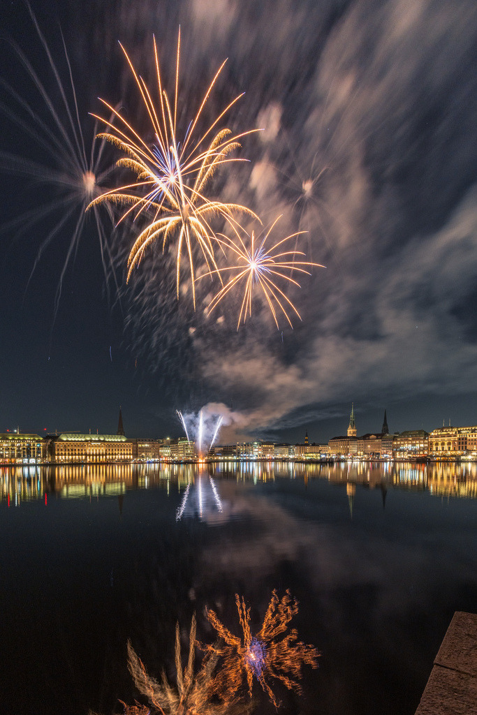 Goldenes Feuerwerk über Hamburg | Goldene Feuerwerksbahnen zeichnen sich vor dem dunklen Hamburger Nachthimmel ab. Die beleuchtete Uferbebauung und ihre Reflexion verleihen der Szene zusätzliche Tiefe. - Realisiert mit Pictrs.com