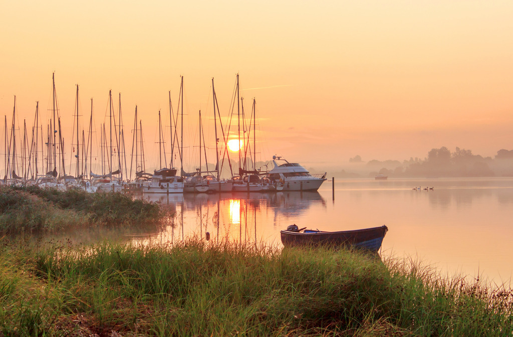 Wandbild: Sonnenaufgang am Ostseefjord | Dieses Wandbild im Querformat zeigt einen schönen Sonnenaufgang am Yachthafen. Im Vordergrund ist Schilf zu sehen. Auf der Schlei ist direkt hinter dem Schilf ein Boot zu sehen. Der Himmel leuchtet in einem schönen dezenten rot. - Realisiert mit Pictrs.com