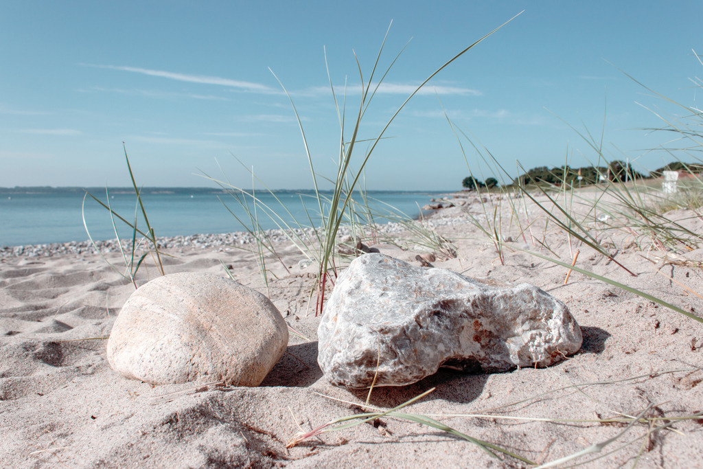Wandbild: Strandgras und Steine am Sandstrand | Dieses Wandbild zeigt Strandgras und zwei Steine am Sandstrand. Das Meer und der Himmel sind pastellartig blau.  Holen Sie sich dieses traumhafte Strandmotiv in dezenten Farben auf Leinwand, auf Aluminium-Platte oder Acrylglas. Ideal fürs Wohnzimmer, Schlafzimmer, Küche, den Arbeitsplatz oder die Ferienwohnung. Die Wandbilder werden individuell für Sie in vielen Abmessungen produziert. Daher passen die Ostseekult Wandbilder immer perfekt an Ihre Wände.  - Realisiert mit Pictrs.com