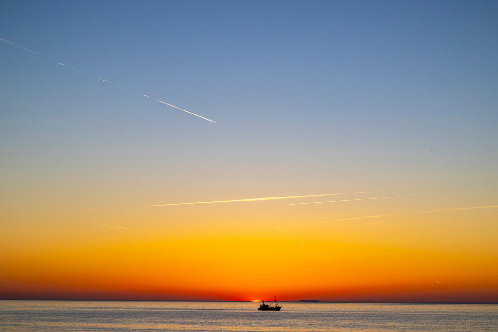 Sonnenuntergang am Meer | Das Foto fängt einen stimmungsvollen Moment auf dem Meer ein. Ein Fischkutter ist als dunkle Silhouette vor einem intensiven Sonnenuntergang zu sehen. Die Sonne ist fast vollständig hinter dem Horizont verschwunden und hinterlässt einen leuchtend orange-gelben Farbverlauf am Himmel, der sich sanft im ruhigen Wasser spiegelt. - Realisiert mit Pictrs.com