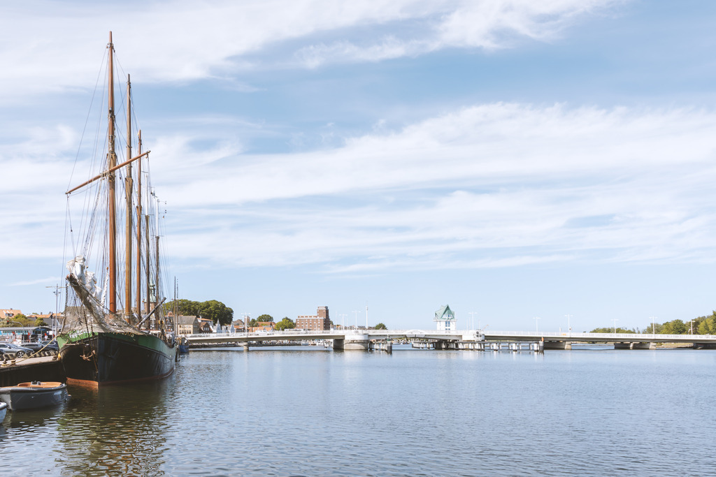 Wandbild: Segelschiff an der Schleibrücke – Maritimes Licht aus Kappeln | Dieses Wandbild zeigt ein Segelschiff vor der Schleibrücke in Kappeln, eingefangen in einem hellen, klaren Licht, das die weite Wasserfläche und die freundliche Atmosphäre der Schlei betont. Die hohen Masten, das ruhige Wasser und der offene Himmel verleihen dem Motiv eine maritime Leichtigkeit. Die Brücke mit ihrem markanten Turm setzt einen architektonischen Akzent, während die umliegenden Gebäude sich harmonisch in die Szenerie einfügen. Das Bild strahlt Ruhe, Weite und norddeutsches Flair aus – ideal für helle Wohnräume, Flure oder Arbeitsbereiche.
