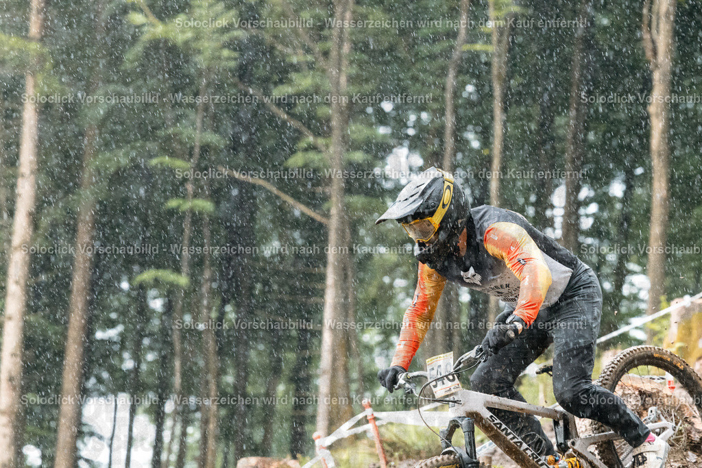 Abenteuer im Regen: Mountainbiker in Aktion | Das Bild zeigt einen Mountainbiker in voller Montur, der einen steilen, bewaldeten Hang hinabfährt. Der Fahrer trägt einen orangefarbenen Helm mit einer Visier, eine dunkle Jacke und Handschuhe. Er fährt ein Mountainbike der Marke Santa Cruz. Der Hintergrund besteht aus dichtem Grünwerk und einem bewaldeten Hang. Es regnet stark, was die Szene dynamisch und herausfordernd erscheinen lässt. - Realisiert mit Pictrs.com