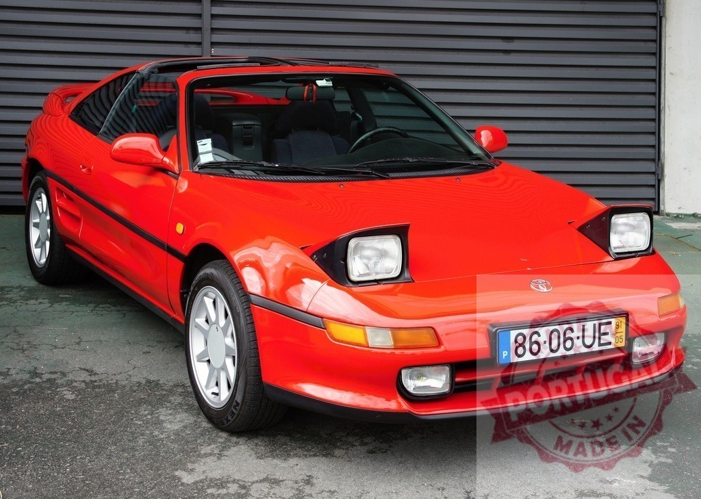 Red Toyota MR2 parked near a gray metallic roll-up door | Gleaming red Toyota MR2 sports car parked near a gray metallic roll-up door, showcasing its sleek design and vibrant color - Realized with Pictrs.com