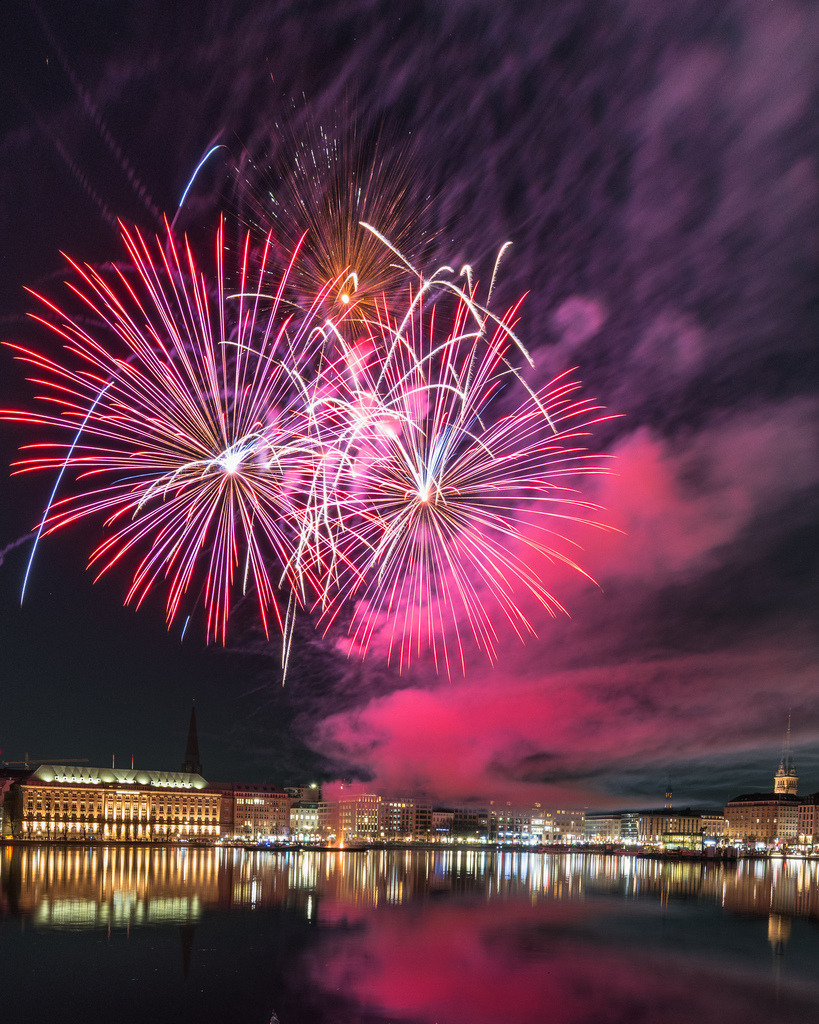 Rotes Feuerwerk über der Hamburger Skyline | Große rote und weiße Feuerwerkssterne dominieren den Himmel über der Alster. Die beleuchtete Hamburger Innenstadt bildet darunter eine ruhige Gegenkulisse. - Realisiert mit Pictrs.com