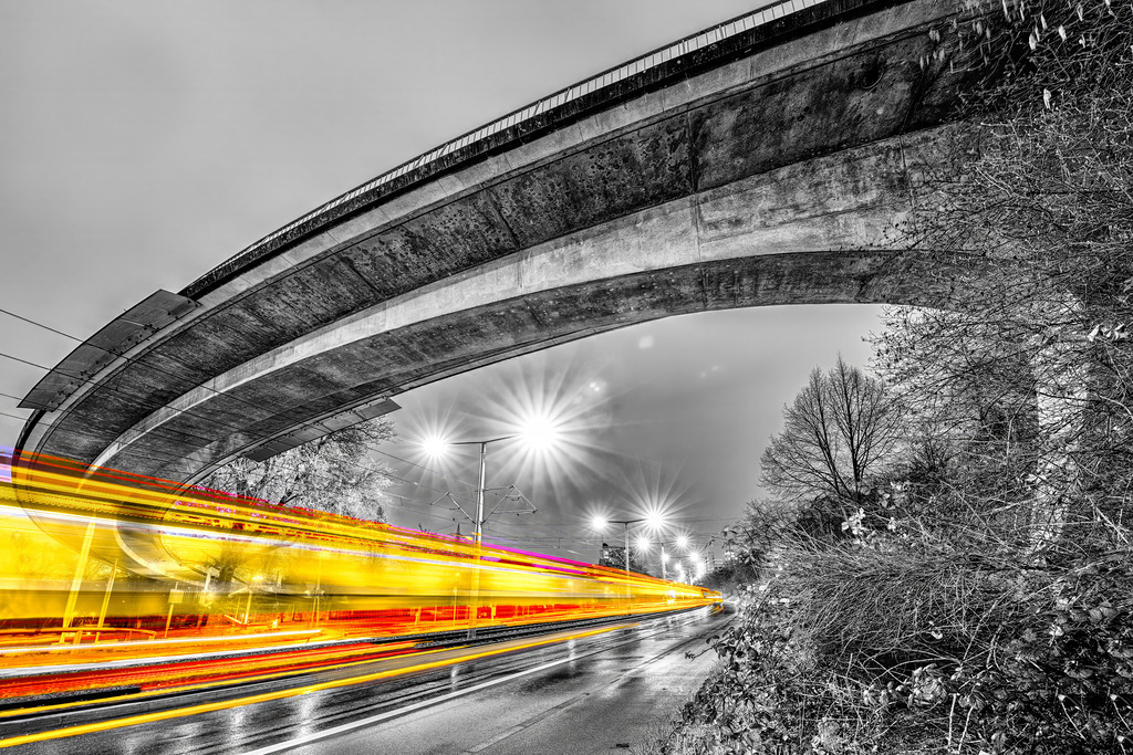 Der La-Ferté-Steg - Stuttgart Zuffenhausen, durchfahren von einer gelben Lichtspur in Schwarz Weiß | Das Bild zeigt eine nächtliche Langzeitbelichtung von Lichtspuren des La-Ferté-Steg in Stuttgart Zuffenhausen, fotografiert von Tom-Pic-Art.Motiv: Eine geschwungene Betonbrücke mit darüber verlaufender Fahrbahn und straßenseitigen Lichtspuren (gelb/rot) von Fahrzeugen im unteren Bildbereich.Schwarz-Weiß-Fotografie (Monochrom) mit selektiver Farbgebung (Color-Key) bei den gelben und roten Lichtstreifen - Realisiert mit Pictrs.com