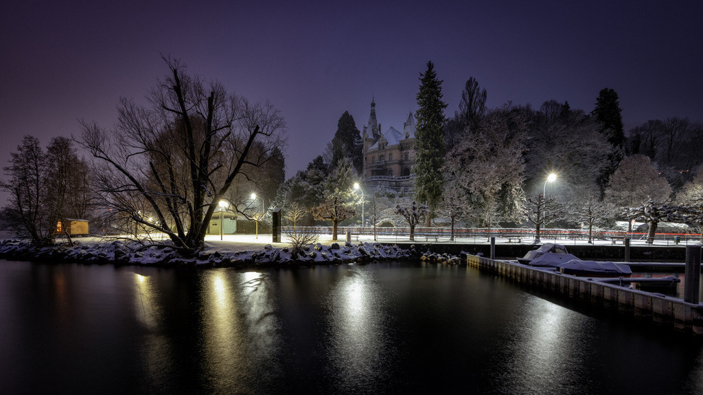 Schloss Hünegg Winternacht am Thunersee | Ein winterliches Panorama zeigt Schloss Hünegg am Ufer des Thunersees in Hilterfingen. Die Szene ist von Schnee bedeckt und wird von Strassenlaternen beleuchtet, deren Licht sich auf der ruhigen Wasseroberfläche spiegelt. Rote Lichtspuren eines Fahrzeugs sind auf der Uferstrasse erkennbar. - Realisiert mit Pictrs.com