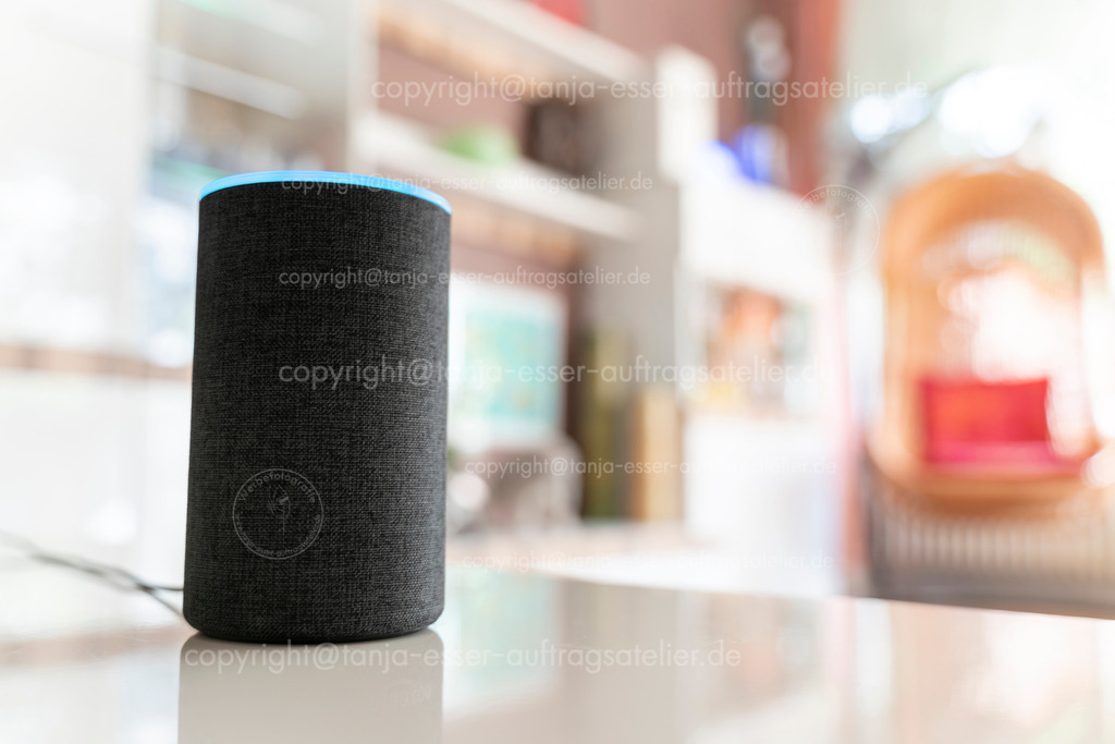 Smarter Lautsprecher in hellem Wohnzimmer D | Ein smarter Lautsprecher Assistent steht in einem hellen Wohnzimmer. Oben leuchtet ein hellblauer Kreis, das Gerät ist auf Empfang. Viel Copy Space.