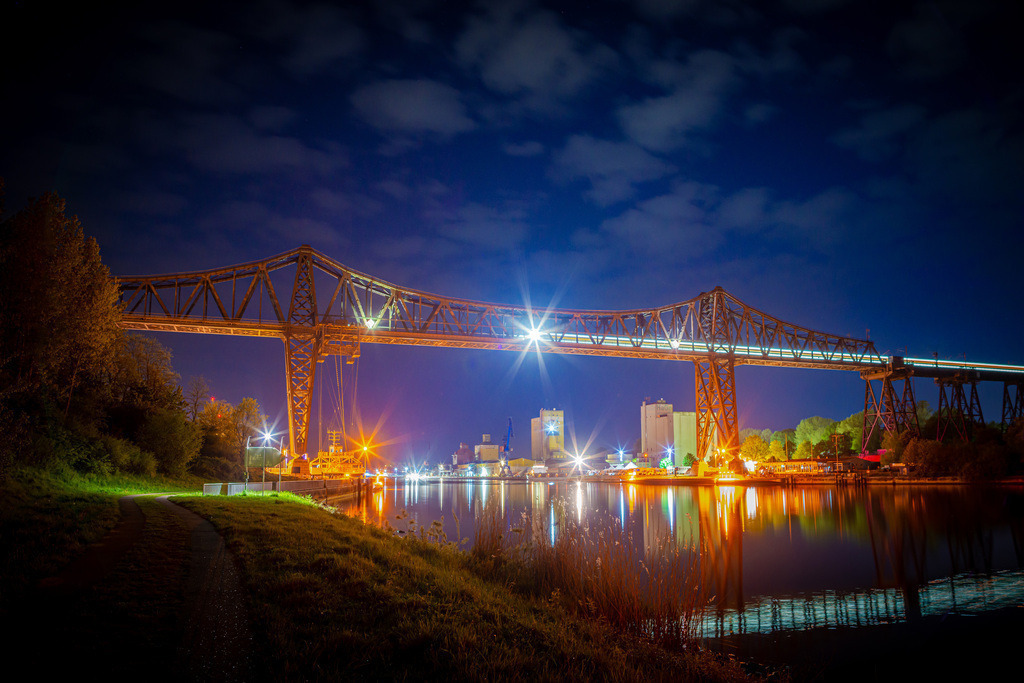 Eisenbahn Hochbrücke Rendsburg | Die Nachtaufnahme zeigt die majestätische Rendsburger Eisenbahn Hochbrücke über dem glitzernden Nord-Ostsee-Kanal. Das warme Licht der Laternen entlang der Brücke und des Kanals schafft eine romantische Stimmung und lässt das Wasser in einem eindrucksvollen Schein erstrahlen. Im Hintergrund erheben sich Kreishafen von Rendsburg. Die Szene wirkt beinahe magisch und lässt den Betrachter in eine verträumte Welt eintauchen.