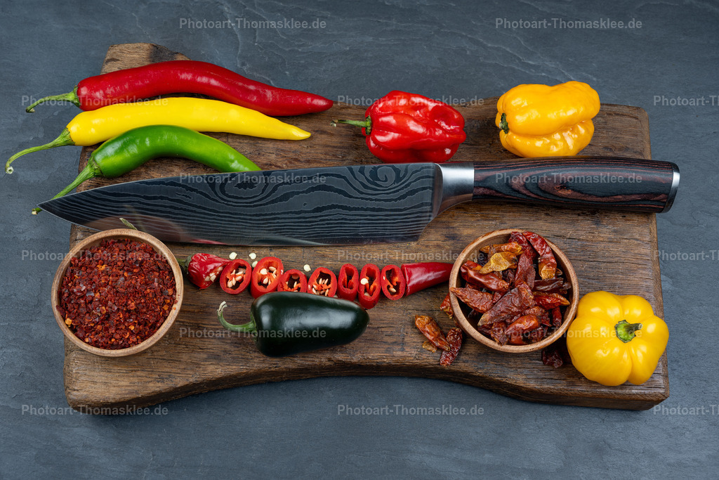 Scharfe Gewürze mit Damastmesser auf Schneidebrett | Auf einem rustikalen Holzschneidebrett arrangieren sich bunte Paprika und Chilis in leuchtenden Farben. Ein elegantes Damastmesser mit dunkler Holzmaserung liegt mittig auf dem Brett. Rote, gelbe und grüne Paprika sowie verschiedene Chilischoten bilden eine harmonische Komposition. Zwei kleine Holzschalen enthalten Chilipulver und getrocknete Chilis. Der dunkle Schieferhintergrund unterstreicht die natürlichen Farben der frischen Gewürze. Diese Küchenszene verkörpert die Kunst des Kochens mit scharfen, aromatischen Zutaten perfekt. - Realisiert mit Pictrs.com