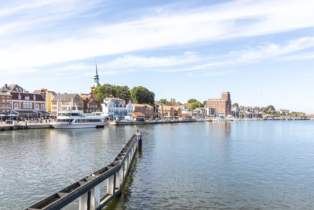 Wandbild: Helles Hafenbild aus Kappeln – Blick auf Kirche und Hafen | Dieses Wandbild zeigt den Hafen von Kappeln in einem klaren, hellen Licht. Die farbigen Gebäude am Wasser, die ruhige Wasserfläche und die markante Kirche mit ihrem grünen Turm prägen die Szenerie und verleihen dem Motiv eine freundliche, maritime Atmosphäre. Das große Schiff im Vordergrund und die Boote im Hafen ergänzen das lebendige, aber dennoch ruhige Gesamtbild. Ein Premium‑Wandbild im klassischen 3:2‑Format, das Weite, Helligkeit und norddeutsches Flair in moderne Wohnräume bringt.
