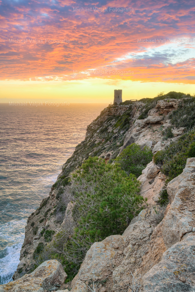 Alter Wachturm am Cap Blanc auf Mallorca | Der Wachturm am Cap Blanc steht auf einer markanten Felsformation, die sich steil über dem Meer erhebt. In der vertikalen Perspektive wird die Höhe der Klippe besonders deutlich, während das gleichmäßige Licht des Sonnenuntergangs die Struktur des Turms und die Schichtung der Felsen klar hervorhebt. Die Szene zeigt die typische Küstenlandschaft Mallorcas in einer ruhigen, sachlichen Abendstimmung. - Realisiert mit Pictrs.com