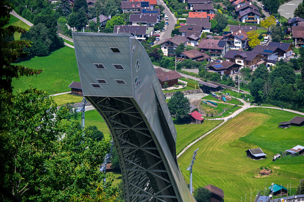 Über den Dächern: Wo Träume fliegen lernen | Ein Anblick, der ehrfürchtiges Staunen und Herzklopfen zugleich auslöst: Majestätisch und kühn ragt die moderne Architektur der Skisprungschanze in Garmisch-Partenkirchen aus einem Meer von sattgrünen Wiesen empor. Wie eine Rampe in den Himmel wirkt ihre elegante, silbrig glänzende Konstruktion mit den markanten Fensterreihen. Weit unten im Tal schmiegen sich die friedlichen Häuser des Ortes mit ihren roten Dächern in die idyllische Alpenlandschaft, bewacht von dunklen Tannenwäldern. Diese Perspektive fängt die dramatische Spannung zwischen menschlichem Wagemut und der erhabenen Ruhe der Berge perfekt ein. Ein Ort, der Sehnsucht nach Freiheit und der Überwindung von Grenzen weckt. - Realisiert mit Pictrs.com