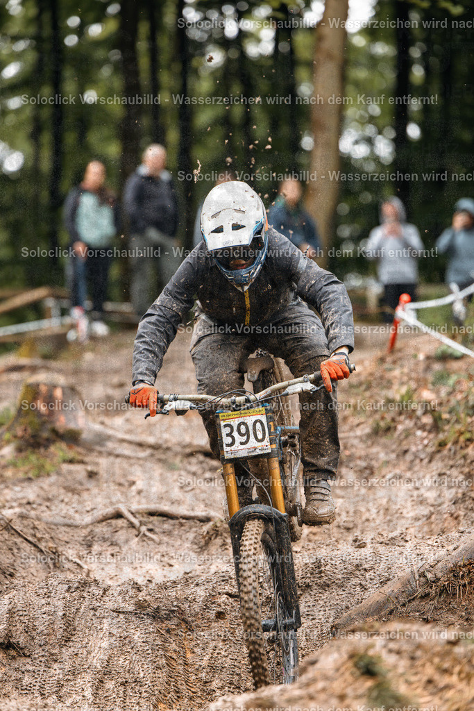 Mountainbike-Action im Wald | Ein Mountainbiker in voller Ausrüstung rast durch einen schlammigen, unebenen Pfad in einem dichten Wald. Er trägt einen weißen Helm mit einer gelben Visier und eine dunkle Schutzkleidung. Der Fokus liegt auf dem Fahrer und seinem Mountainbike, während der Hintergrund verschwommen ist. Der Pfad ist mit Schlamm bedeckt, was die Herausforderung des Geländes unterstreicht. - Realisiert mit Pictrs.com