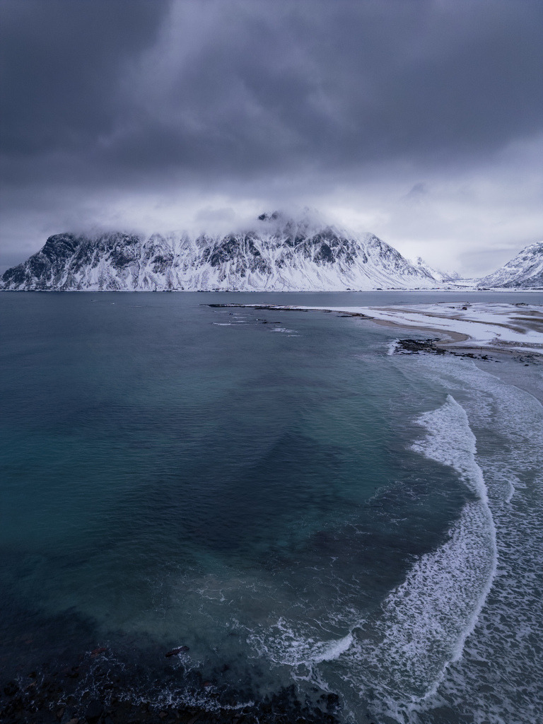 Arktische Küste aus der Vogelperspektive | Dunkles Meer trifft auf eine verschneite norwegische Küstenlinie unter schweren Wolken. Die Luftaufnahme zeigt die raue Struktur der arktischen Landschaft und die starken Kontraste zwischen Wasser, Schnee und Bergen. - Realisiert mit Pictrs.com