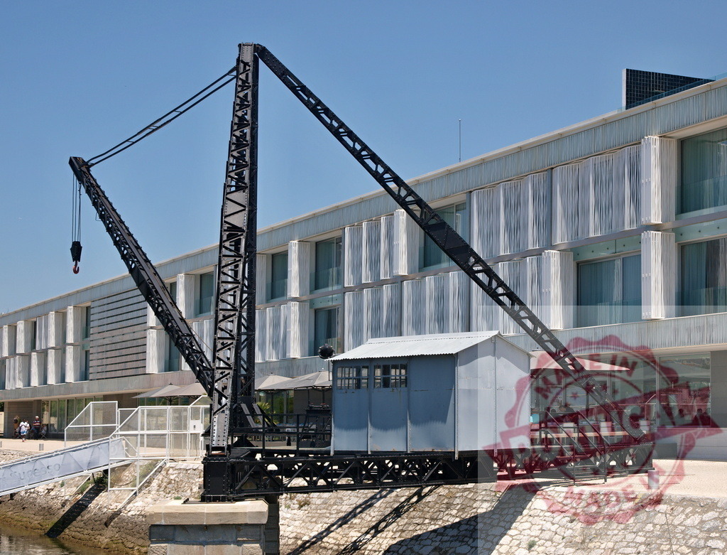 Old industrial crane contrasting modern building architecture. Lisbon - Portugal | Historic harbor crane standing beside contemporary waterfront building - Realisiert mit Pictrs.com