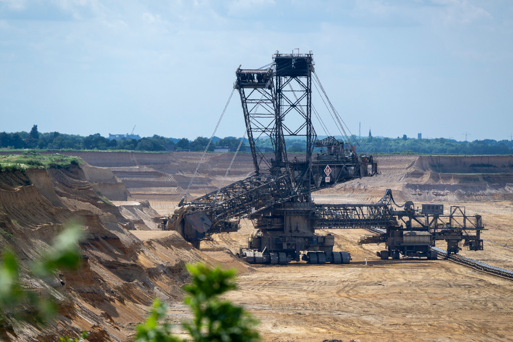 Gigant der Erdbewegung: Schaufelradbagger im Tagebau | Das Foto dokumentiert die gewaltige Dimension des Braunkohletagebaus. Im Zentrum steht ein monumentaler Schaufelradbagger, dessen komplexe Stahlkonstruktion und riesige Ausleger die karge, sandig-braune Abbaugrube dominieren. Während der Vordergrund durch vereinzelte grüne Blätter natürlich eingerahmt wird, steigen im Hintergrund die hellen, terrassenförmigen Erdschichten empor. An der oberen Abbaukante wird die ursprüngliche, grüne Landschaft unter einem hellen Himmel sichtbar. Eine eindrucksvolle Aufnahme industrieller Landschaftsveränderung. - Realisiert mit Pictrs.com
