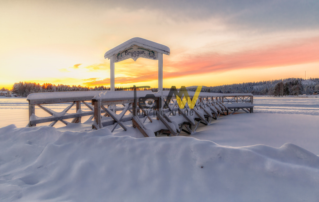 Winterliche Seelandschaft in Schweden|Toller Sonnenaufgang | Eine schneebedeckter Steg, der zu einem Anlegesteg mit einem Schild führt, auf dem "Bryggan" steht. Die Szene ist von Schnee bedeckt und der Himmel zeigt einen Sonnenuntergang in Orange- und Rosatönen. - Realisiert mit Pictrs.com