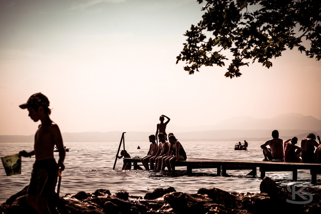 Stimmungsvolle Badeszene am Gardasee bei Sonnenuntergang | Ein idyllisches Bild einer Badeszene am Gardasee. Das warme, weiche Licht des Sonnenuntergangs beleuchtet das Wasser und die umliegenden Berge, während Menschen in Silhouette am Steg sitzen und schwimmen. Die Aufnahme fängt die friedliche und nostalgische Urlaubsatmosphäre perfekt ein. - Realisiert mit Pictrs.com