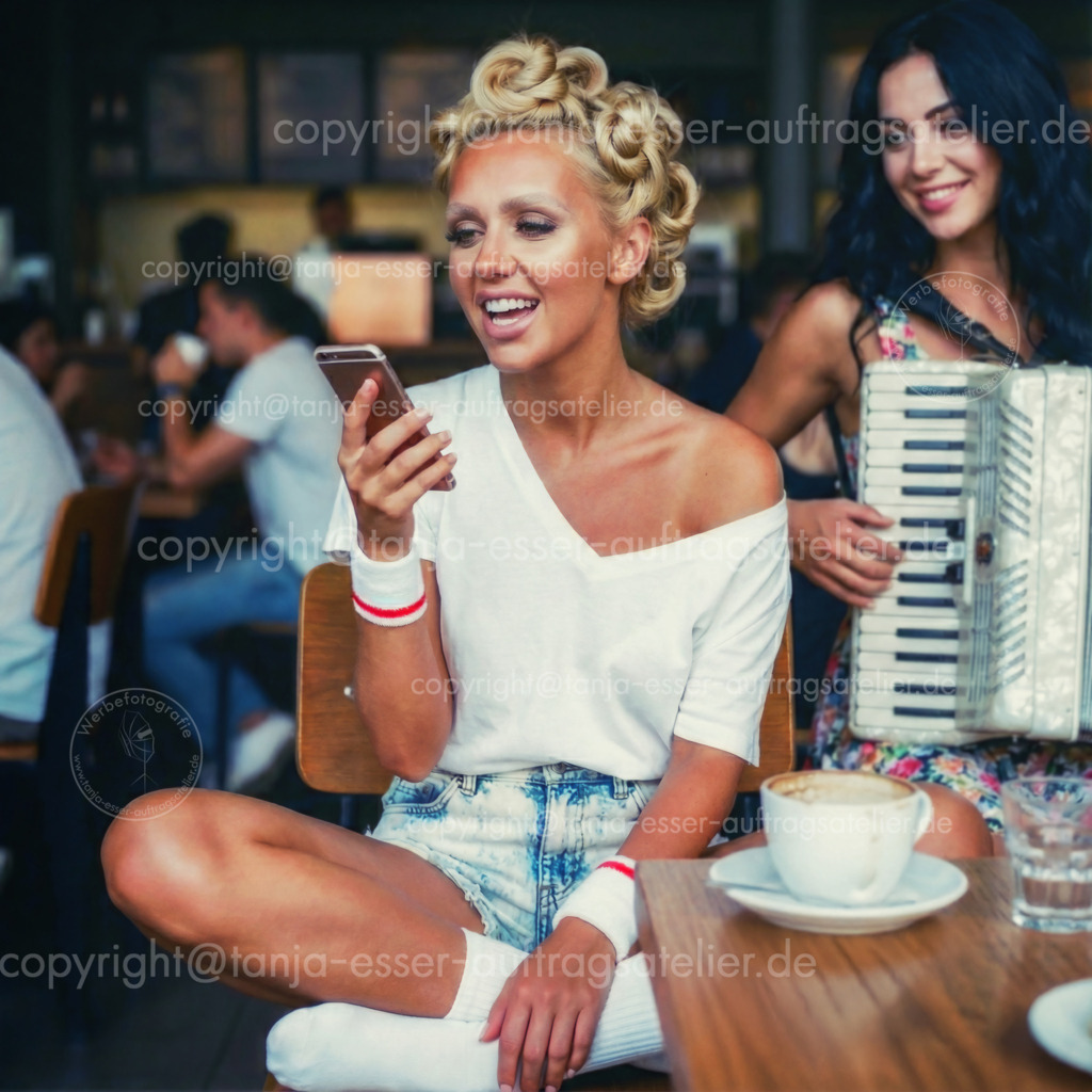 Musik im Coffee Shop | Moderne, entspannte junge Frau singt im Café mit Smartphone, begleitet von junger Akkordeonspielerin – Lässiger Look mit Tennissocken, Schweißbändern und Hochsteckfrisur. Das Bild wurde nach eigenen Vorstellungen von einer KI erstellt und überarbeitet. 
