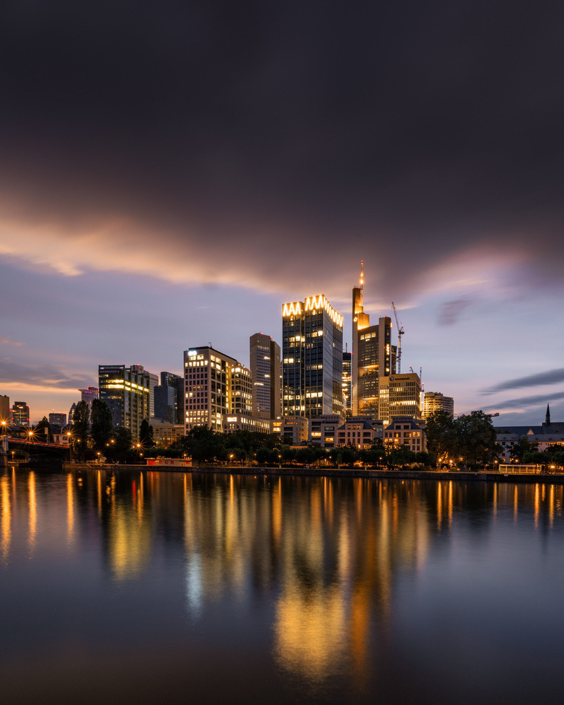 Frankfurter Skyline unter dramatischen Wolken | Die beleuchtete Skyline von Frankfurt erhebt sich am Main unter einem dramatischen Abendhimmel. Die lange Belichtung glättet das Wasser und verstärkt die Spiegelungen der Hochhäuser. - Realisiert mit Pictrs.com