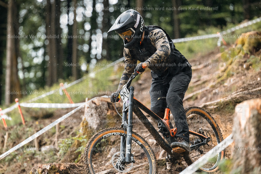 Downhill-Action im Schnee | Ein Mountainbiker in voller Ausrüstung rast einen steilen, verschneiten Hang hinunter. Er trägt einen schwarzen POC-Helm und eine Jacke mit Tarnmuster, sowie schwarze Handschuhe und Hosen. Das Gelände ist mit Bäumen und Büschen bewachsen, und es sind Markierungen für einen Downhill-Kurs zu sehen. Der Fahrer scheint sich voll und ganz auf die Fahrt zu konzentrieren. - Realisiert mit Pictrs.com