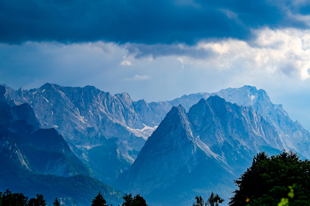 Urgewalt über den Gipfeln: Das Leuchten der Alpen | Ein Anblick von atemberaubender Dramatik und erhabener Schönheit: Unter einer mächtigen, tiefblau grollenden Wolkenwand zeigt sich das gewaltige Wettersteingebirge in seiner rauen, ungezähmten Pracht. Während düstere Schatten über den schroffen Steilwänden und Tälern liegen, bricht plötzlich gleißendes Sonnenlicht durch die Wolkendecke und lässt die markanten Felsgipfel sowie die Zugspitze im Hintergrund magisch erstrahlen. Diese Aufnahme fängt das faszinierende, fast mystische Wechselspiel der Elemente im Hochgebirge ein – ein Bild voller Ehrfurcht, Einsamkeit und unendlicher Sehnsucht nach der Wildnis. - Realisiert mit Pictrs.com