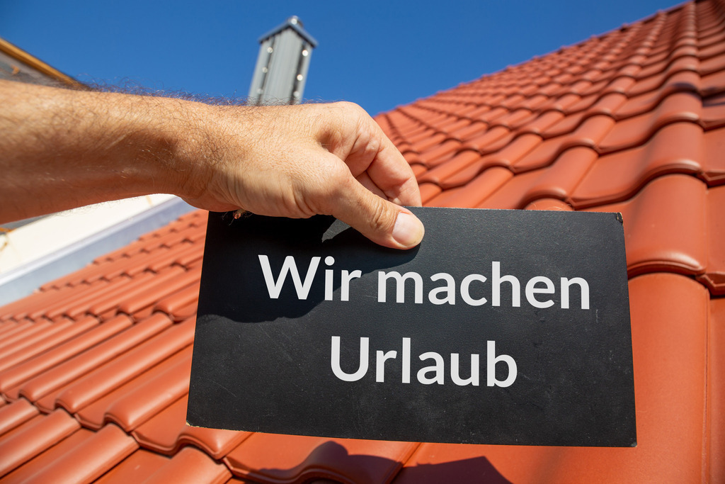 Wir machen Urlaub - Betriebsurlaub Vorlage ideal für Dachdecker | Betriebsferien Dachdecker: Mann hält ein Schild (mit hinzugefügtem Text) WIR MACHEN URLAUB vor einem neuen Ziegeldach - Realisiert mit Pictrs.com