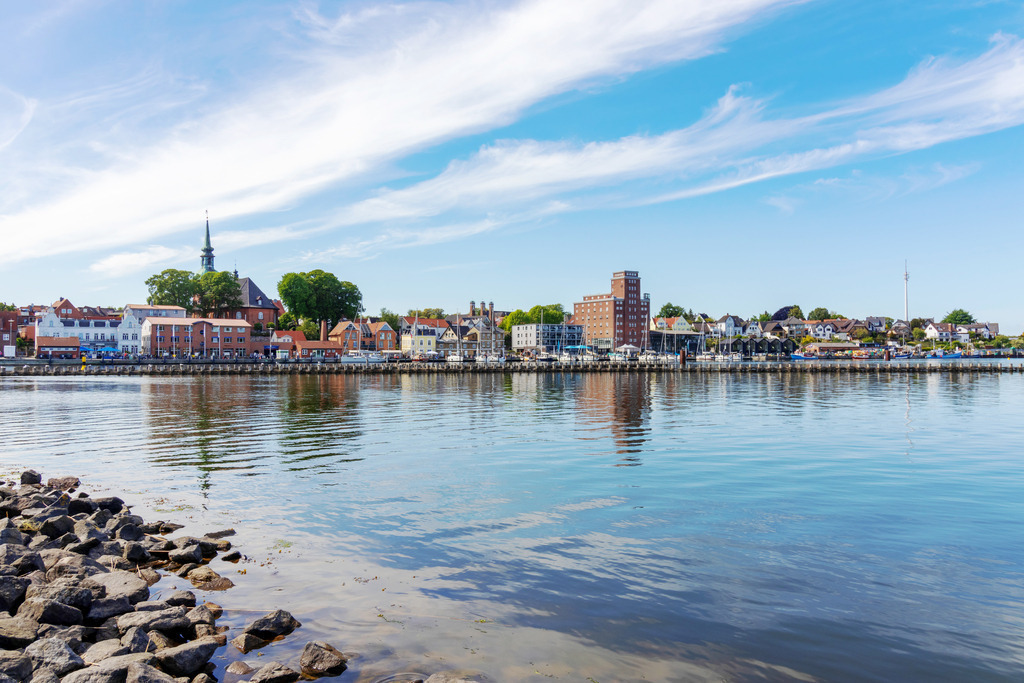 Wandbild: Spiegelung am Hafen in Kappeln an der Schlei | Dieses Wandbild im Querformat zeigt den Hafen in Kappeln an der Schlei. Auf dem ruhigen Wasser spiegeln sich die Gebäude auf der Schlei. Im Vordergrund auf der linken Seite befinden sich Steine zur Uferbefestigung. Am blauen Himmel sind einige Schleierwolken zu sehen.  - Realisiert mit Pictrs.com