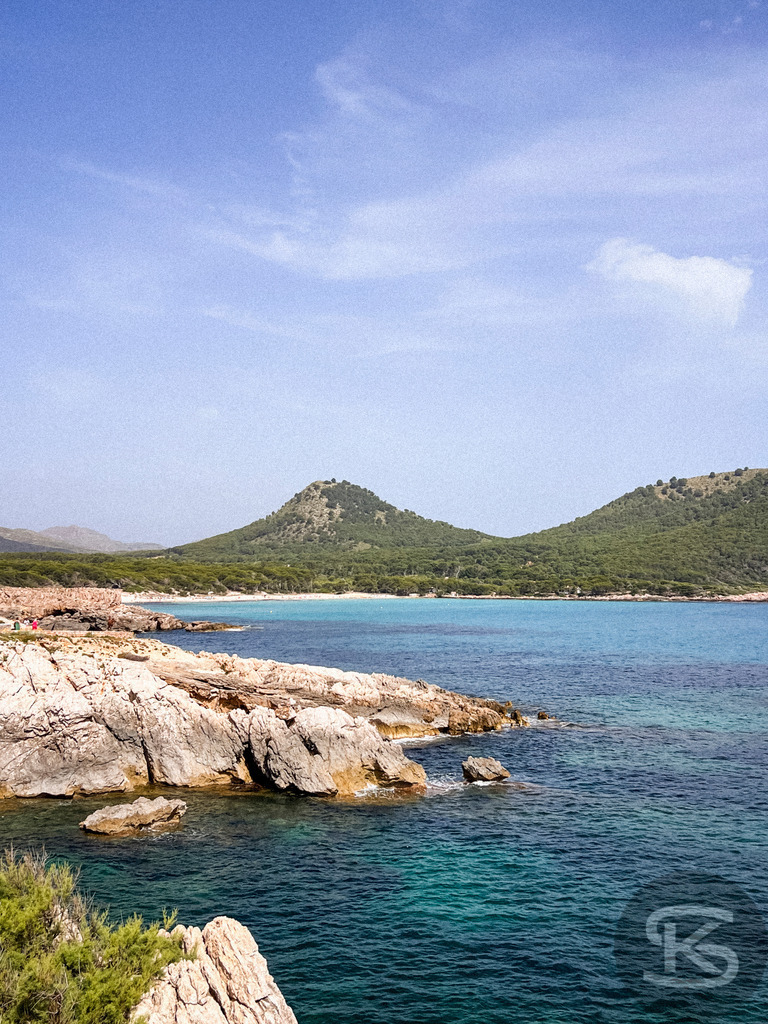 Blick über Felsen auf die Bucht der Cala Agulla und den Berg Es Telégraf, Mallorca | Vordergrund mit hellen Küstenfelsen, dahinter die türkisfarbene Bucht Cala Agulla und ein bewaldeter, kegelförmiger Berg unter blauem Himmel auf Mallorca. - Realisiert mit Pictrs.com