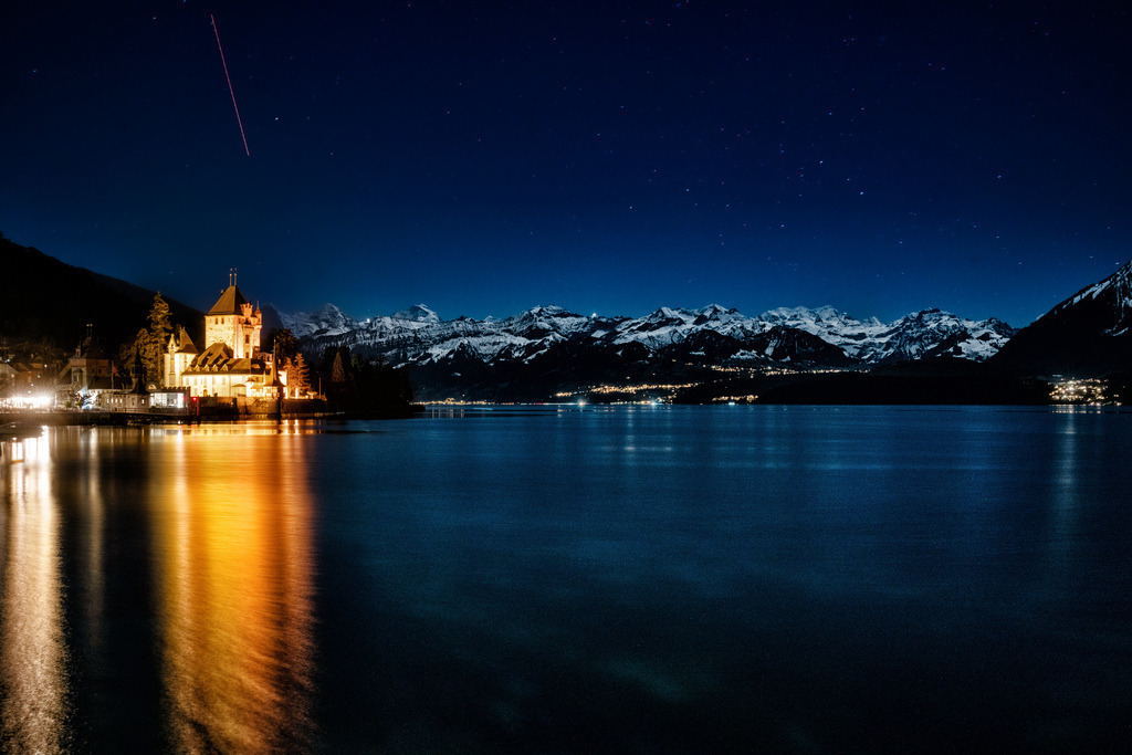 Alpenmagie | Eine weite Nachtlandschaft zeigt ein beleuchtetes Schloss am Ufer eines Sees, umgeben von schneebedeckten Bergen unter einem sternenklaren Himmel. Links dominiert das Schloss Oberhofen mit seinen warm leuchtenden Mauern und Türmen, deren goldenes Licht sich lang und streifenförmig im dunklen, leicht bewegten Seewasser spiegelt. Rechts vom Schloss erstreckt sich der See in tiefem Blau, mit weiteren fernen Lichtern am gegenüberliegenden Ufer, die ebenfalls schwach reflektiert werden. Im Hintergrund ragt eine Kette majestätischer, schneebedeckter Berggipfel empor, die sich dunkel vom tiefblauen Nachthimmel abheben. Der Himmel ist übersät mit unzähligen kleinen Sternen, und diagonal im oberen linken Bereich zieht sich ein feiner, roter Lichtstreifen durch die Dunkelheit. Die Komposition ist ruhig und weitläufig, mit einem starken Kontrast zwischen dem warmen Licht des Schlosses und den kühlen Blautönen der Nacht. - Realisiert mit Pictrs.com
