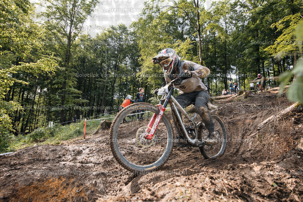 Downhill-Action im Wald | Ein Actionfoto eines Mountainbikers, der einen Downhill-Kurs bewältigt. Der Fahrer ist in voller Montur und fährt mit hoher Geschwindigkeit einen steilen, mit Wurzeln und Schlamm bedeckten Hang hinunter. - Realisiert mit Pictrs.com