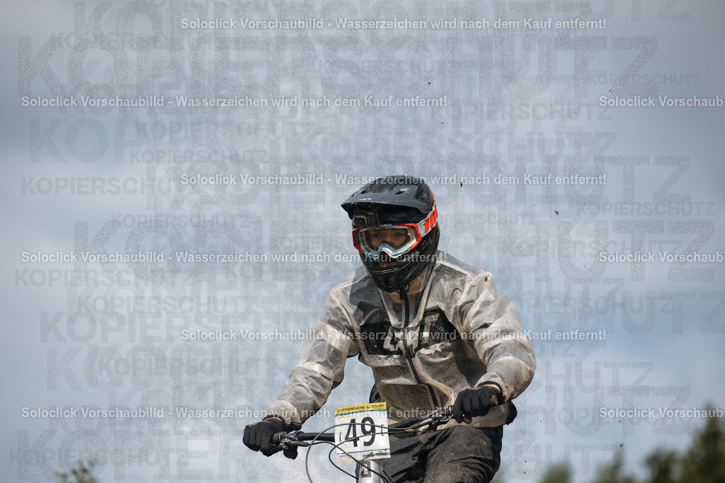 Mountainbike-Action | Ein Mountainbiker in voller Ausrüstung rast auf einem staubigen Pfad. Er trägt einen schwarzen Helm mit Visier, eine Schutzbrille, eine Jacke und Handschuhe. Er hält ein Mountainbike mit breiten Reifen und einer Federgabel. Der Hintergrund besteht aus Bäumen und einem bewölkten Himmel. - Realisiert mit Pictrs.com