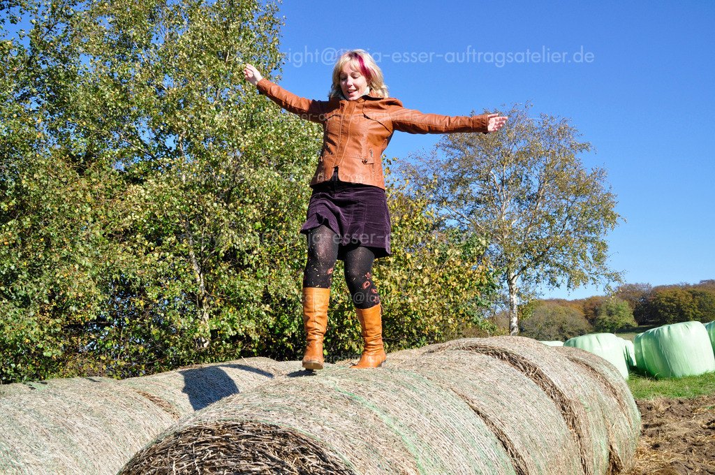 Autumn fun  | Herbstspaß. Eine blonde Frau Ende 30 klettert im schicken Outfit über Siloballen. Lederjacke und Lederstiefel. Sie hat blonde Haare und eine pinke Haarsträhne. Sie lacht und balanciert. 
