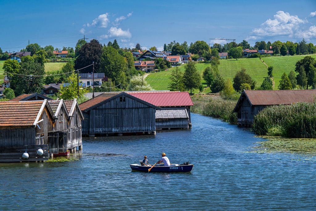 Sommerliche Flussidylle in Seehausen: Bootstour durch die Reethütten | in malerisches Bild oberbayerischer Gemütlichkeit: Ein kleineres Ruderboot gleitet sanft durch die Wasserstraße zwischen urigen, wettergegerbten Holzboothäusern in Seehausen am Staffelsee. Ein Mann mit Strohhut und sein Begleiter genießen die ruhige Fahrt entlang des reetbewachsenen Ufers. Im Hintergrund erstrecken sich saftig grüne Wiesen, friedliche Wohnhäuser und sanfte Hügel unter strahlend blauem Sommerhimmel. Eine herzerwärmende Aufnahme, die Urlaubsstimmung, Tradition und die unbeschwerte Schönheit des Blauen Landes spürbar einfängt. - Realisiert mit Pictrs.com
