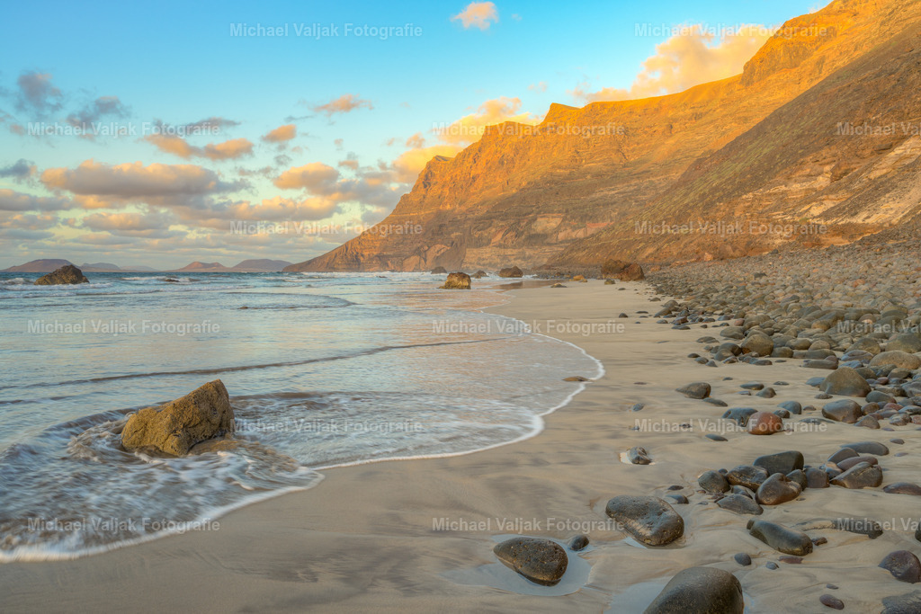 Abendstimmung am Playa de Famara auf Lanzarote | Wenn die Sonne hinter Lanzarote versinkt, verwandelt sich der Playa de Famara in ein Schauspiel aus Licht und Farbe. Die gewaltigen Klippen leuchten im warmen Rot des Abendhimmels, ihre Konturen scharf hervorgehoben durch das letzte Strahlen des Tages. Der weite Strand liegt ruhig zu Füßen dieser Kulisse, während Meer und Himmel sanft ineinander übergehen. Ein Moment, der die wilde Schönheit der Nordküste in eindrucksvoller Klarheit offenbart. - Realisiert mit Pictrs.com