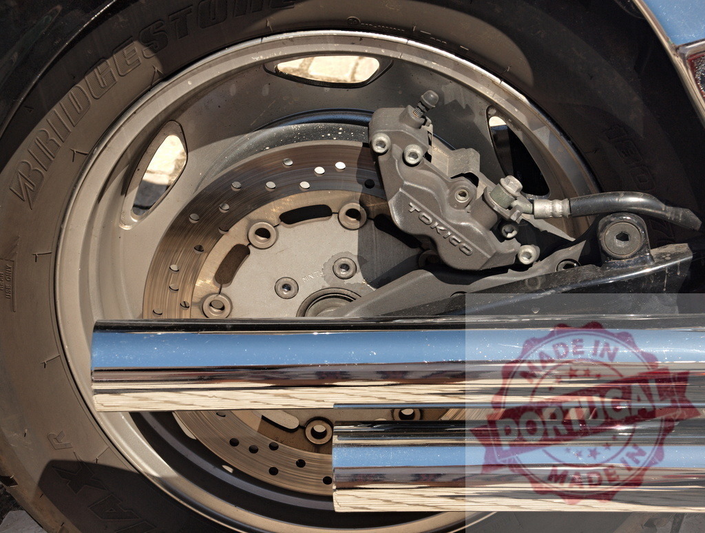 Motorcycle wheel showing disc brake and chrome exhaust pipe | Motorcycle rear wheel featuring a Tokico brake caliper, Bridgestone tire, and shining chrome exhaust - Realized with Pictrs.com