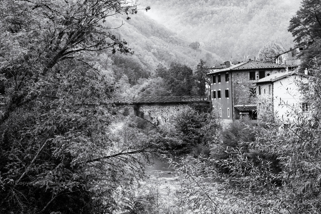 Haus an Steinbrücke in Bagni di Lucca Toskana | Bagni di Lucca ist ein bezaubernder Ort in der Region Lucca Toskana. Die Stadt am Flusslauf des Lima liegt in einer beeindruckenden Berglandschaft.
Schwarz weiß Fotografie eines typisch toskanischen Hauses mit Steinbrücke über den Fluss. Im Hintergrund die Berglandschaft, durch die Nebel zieht. - Realisiert mit Pictrs.com