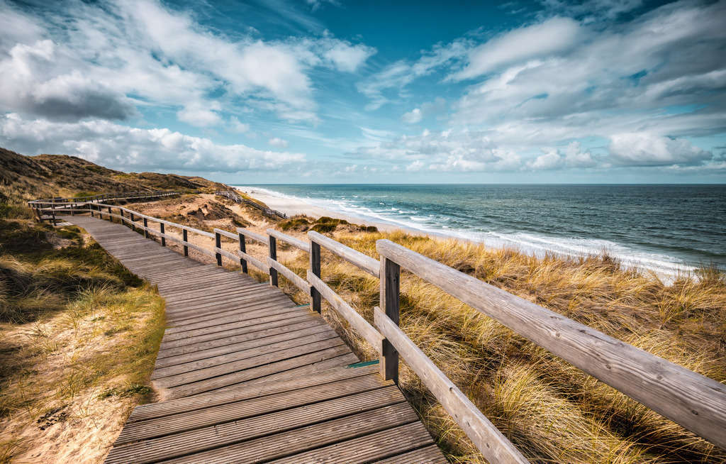 Holzsteg durch die Dünen zum Strand am Meer entlang,Sylt,Nordsee | Das Bild zeigt einen hölzernen Steg durch die Dünen am Strand von Wenningstedt auf der Nordseeinsel Sylt.Wenningstedt-Braderup auf Sylt, Schleswig-Holstein.Typische Dünenlandschaft mit Strandhafer an der Nordseeküste.Wege: Gut ausgebaute Holzstege schützen die empfindlichen Dünen und führen die Besucher direkt zum Sandstrand - Realisiert mit Pictrs.com