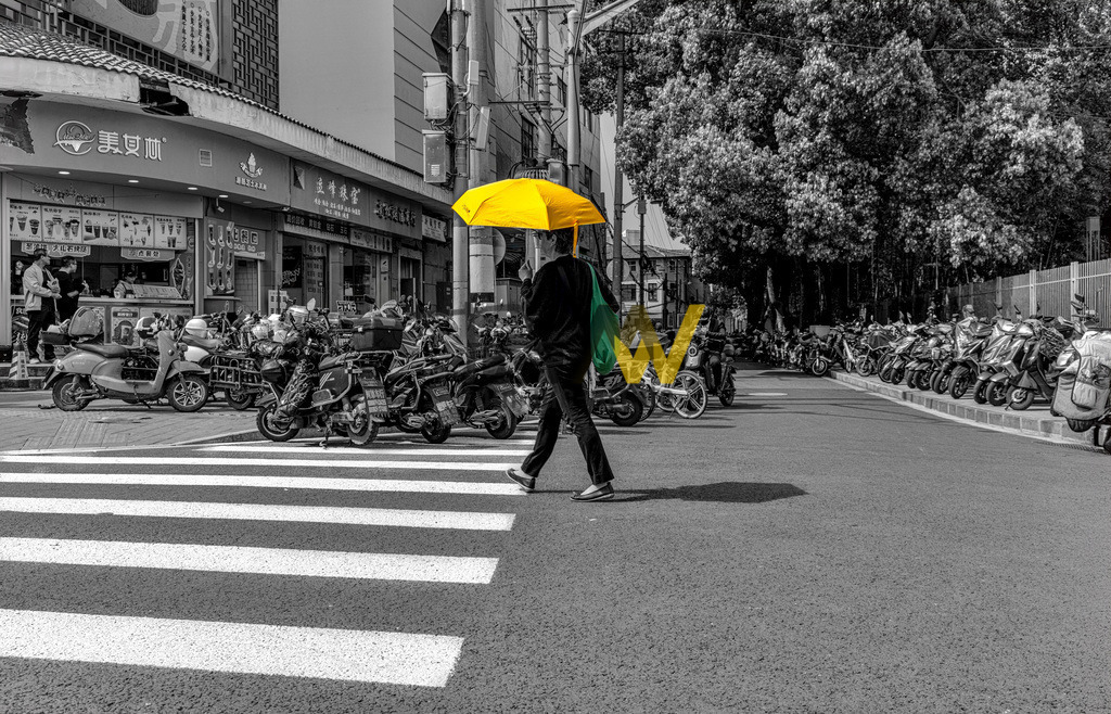 Eine Person mit einem gelben Schirm überquert eine Straße in Shanghai | Das Bild zeigt einen Moment in einer belebten Umgebung in Shanghai, der durch selektive Kolorierung hervorgehoben wird.Eine Person mit einem leuchtend gelben Regenschirm und einer grünen Tasche überquert einen Zebrastreifen.Die Person ist der einzige farbige Akzent in einer ansonsten schwarz-weiß gehaltenen Szene.Zahlreiche geparkte Motorroller und Fahrräder säumen die Straße und den Bürgersteig. - Realisiert mit Pictrs.com