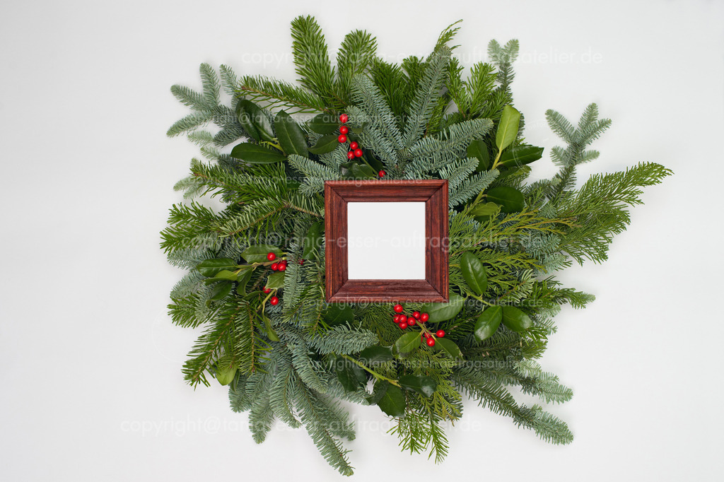 Picture frame with german text:  Merry Christmas in front of fir green | Ein Kreis aus Zweigen verschiedener Tannensorten. Grüne und blaue Tannen. Ein brauner Rahmen aus Holz mit Copy Space. Mock Up.
