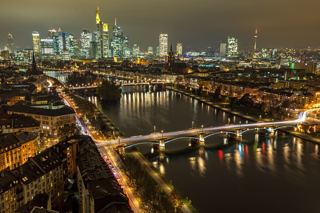 Wandbild - Frankfurts Glanz: Die Strahlende Skyline bei Nacht | Dieses faszinierende Bild zeigt die beleuchtete Skyline von Frankfurt am Main bei Nacht, ein eindrucksvolles Beispiel für die moderne Urbanität der Stadt. Die glitzernden Wolkenkratzer, darunter das ikonische Commerzbank-Hochhaus, dominieren den Horizont und spiegeln sich im ruhigen Wasser des Mains wider. Die zahlreichen Brücken, die über den Fluss führen, sind ebenfalls beleuchtet und tragen zum lebendigen Stadtbild bei. Die langen Lichtspuren der Autos auf den Straßen fügen der Szene eine dynamische Note hinzu. Die Mischung aus historischen und modernen Gebäuden verleiht der Stadt ein einzigartiges Flair, das in diesem Bild wunderschön zur Geltung kommt.