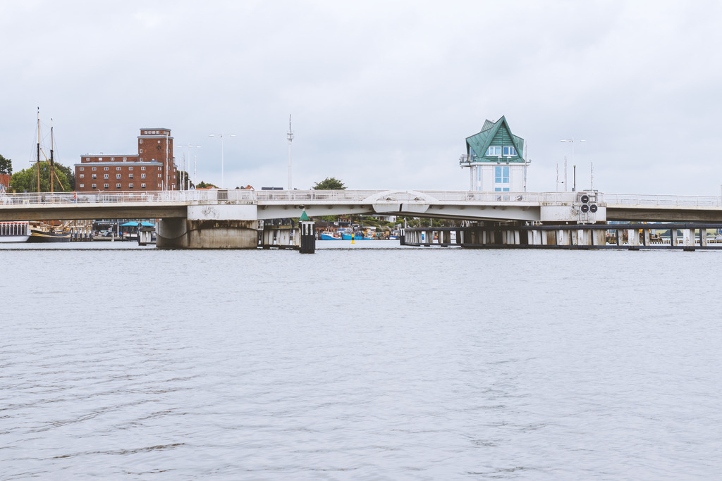 Weite an der Schlei – Die Brücke im norddeutschen Licht | Dieses Wandbild zeigt die Klappbrücke von Kappeln in einem besonders hellen, klaren Licht. Die weite Wasserfläche, der zurückhaltende Himmel und die sanften Farbtöne verleihen dem Motiv eine ruhige, norddeutsche Atmosphäre. Der markante Brückenturm setzt einen feinen architektonischen Akzent, ohne die Leichtigkeit der Szene zu stören. Im Hintergrund liegen historische Gebäude und maritime Details, die sich harmonisch in das helle Gesamtbild einfügen. Dieses Wandbild wirkt bewusst reduziert, freundlich und weit – ideal für Räume, die Ruhe, Klarheit und nordisches Flair ausstrahlen sollen.