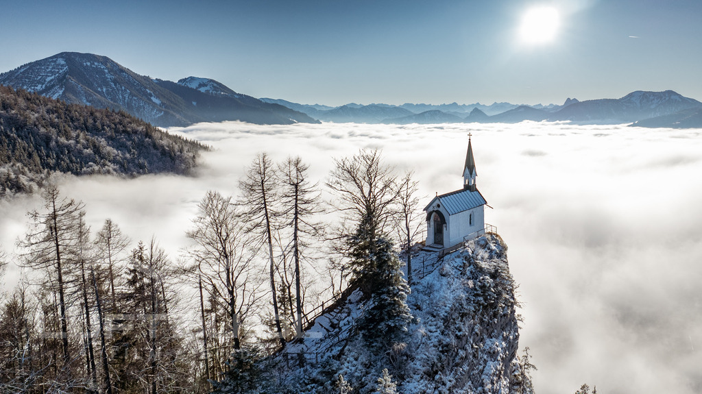 Riederstein über den Wolken; W34 | Der Riederstein erhebt sich hier eindrucksvoll über einem Meer aus Wolken: Die kleine Kapelle thront ruhig auf dem schneebedeckten Felsen, während sich darunter die dichte Wolkendecke wie ein weiches Tuch über das Tal legt. In der klaren Winterluft entsteht ein fast schwebender Eindruck – ein besonderer Moment zwischen Himmel und Erde, der die Magie des Tegernseer Tals spürbar macht. - Realisiert mit Pictrs.com