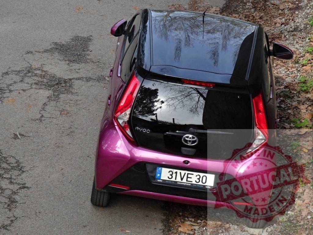 Toyota Aygo compact car parking on asphalt street | Purple Toyota Aygo parked on a cracked paved road next to dry leaves and plants - Realized with Pictrs.com