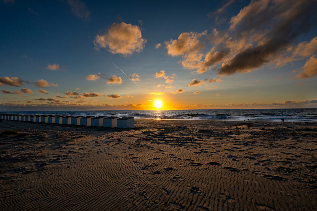 Weiße Badehäuschen säumen die Dünenlandschaft | Reihe traditioneller weißer Strandhäuschen am breiten Sandstrand von Texel während des Sonnenuntergangs über der Nordsee. Die tief stehende Sonne taucht die lockere Bewölkung und die Küstenlandschaft der niederländischen Wattinsel in warmes, goldensequentes Licht. Im Vordergrund zeichnen sich Fußspuren und Strukturen im Sand ab. Redaktionell vielseitig einsetzbar für Berichte über Tourismus, Nordseeurlaub, Reiseziele in den Niederlanden und Entspannung an der Küste. - Realisiert mit Pictrs.com