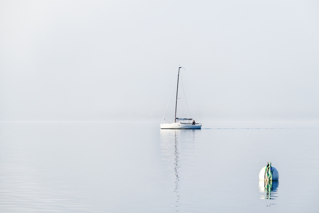 Segelboot auf Thunersee, Hilterfingen | Ein weisses Segelboot mit einer Person an Bord bewegt sich auf spiegelglattem Wasser unter einem hellen Himmel. Die minimalistische Komposition betont die Weite und Ruhe der Szene, mit einer Boje im Vordergrund rechts, die Tiefe verleiht. Das sanfte Licht des Vormittags unterstreicht die friedliche Atmosphäre. - Realisiert mit Pictrs.com