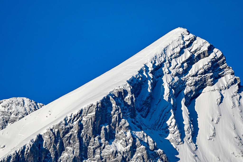 Dem Himmel so nah – Die Alpspitze in voller Pracht | Ein majestätischer Gipfel, der stolz in das tiefe, makellose Blau des Himmels ragt: Die markante Alpspitze. Diese Aufnahme fängt die unberührte, raue Schönheit der winterlichen Bergwelt im berühmten Zugspitzgebiet bei Garmisch-Partenkirchen ein. Während sich schroffe Felswände im Schatten verbergen, strahlt die schneebedeckte Flanke im hellen Sonnenlicht. Ganz oben trotzt das Gipfelkreuz den Elementen – ein Symbol der Sehnsucht, Freiheit und des Triumphs. Ein Bild, das die erhabene Stille der Alpen spürbar macht. - Realisiert mit Pictrs.com